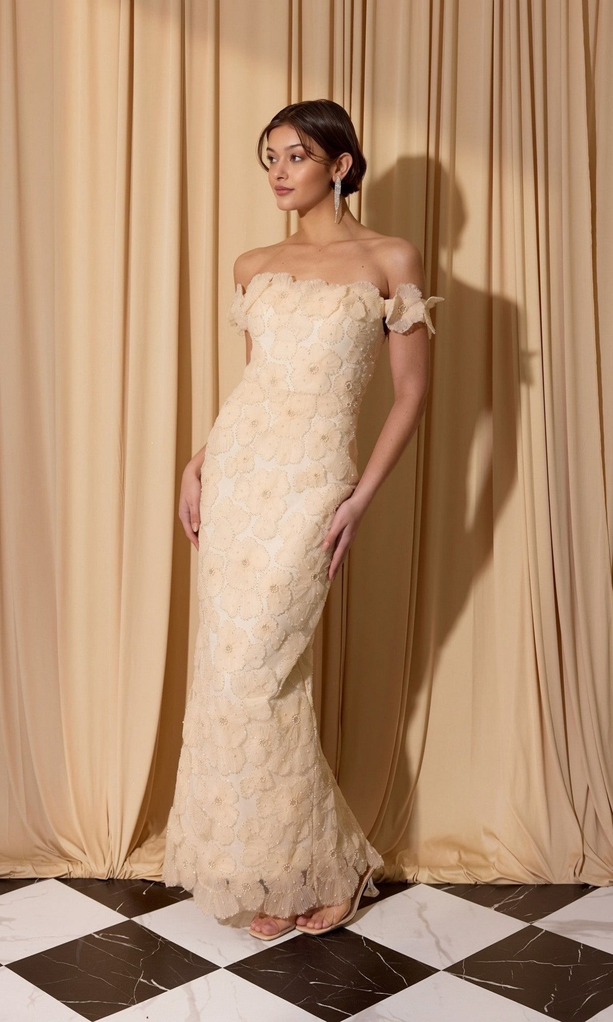 A woman stands on a black and white checkered floor in front of beige curtains, wearing the Eureka EK205 Long Prom Dress in cream, its elegant off-the-shoulder lace design flowing gracefully to the floor.